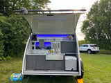 Compact RV rental teardrop trailer shows its fully equipped mobile kitchen with blue lights, perfect for camping.