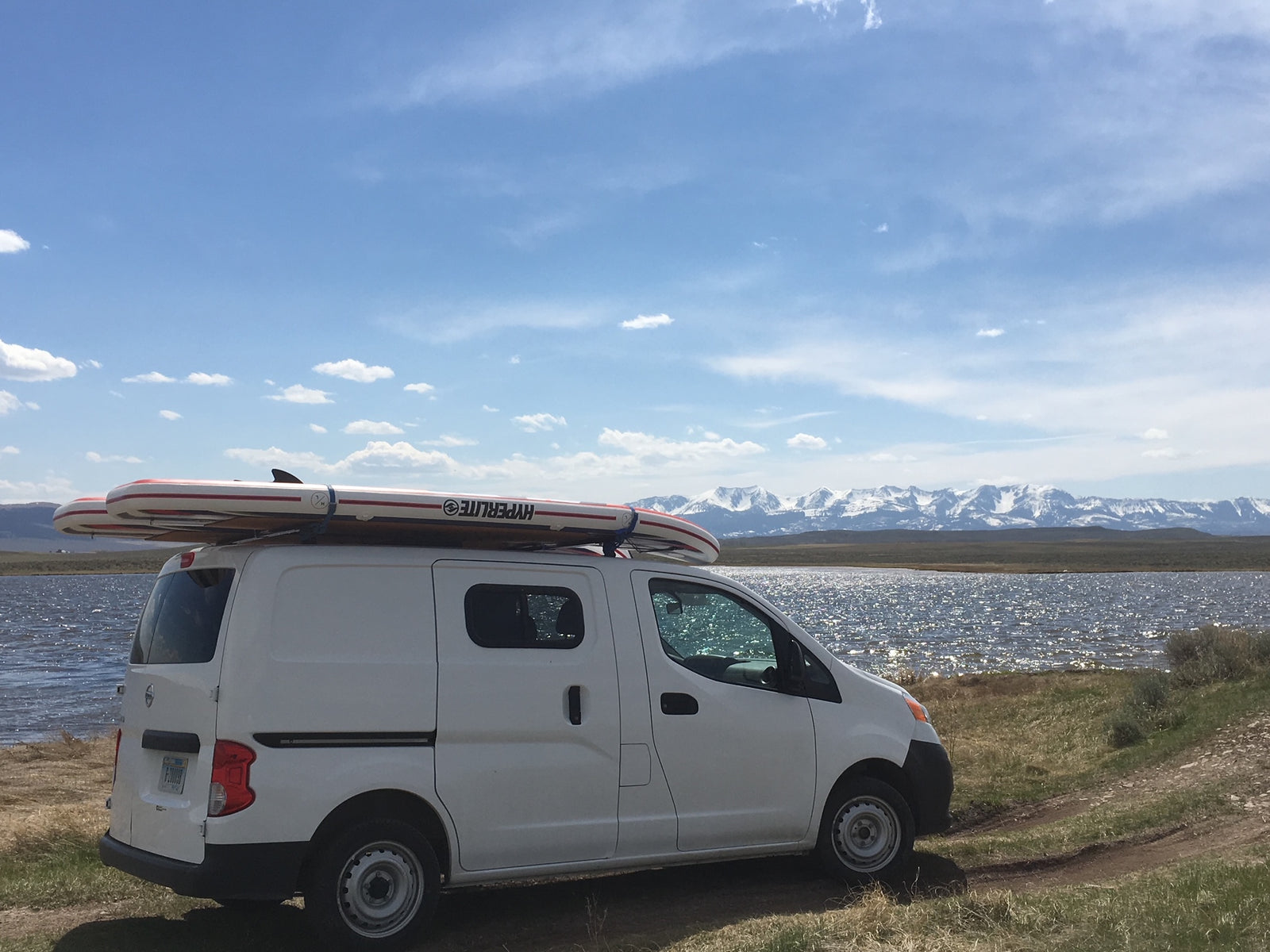 White van with paddleboards on roof by a sparkling lake, snow-capped mountains in distance. Great rv rental for adventure!