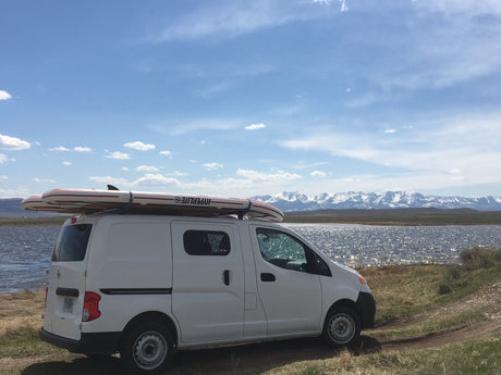 White van with paddleboards on roof by a sparkling lake, snow-capped mountains in distance. Great rv rental for adventure!