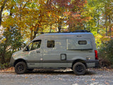 Gray Winnebago Revel 4x4 camper van on a gravel path, surrounded by vibrant autumn trees. Perfect RV rental.