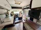 View inside a modern rv rental showing a kitchen counter with sink, swivel front seats, overhead storage, and a lounge with TV.