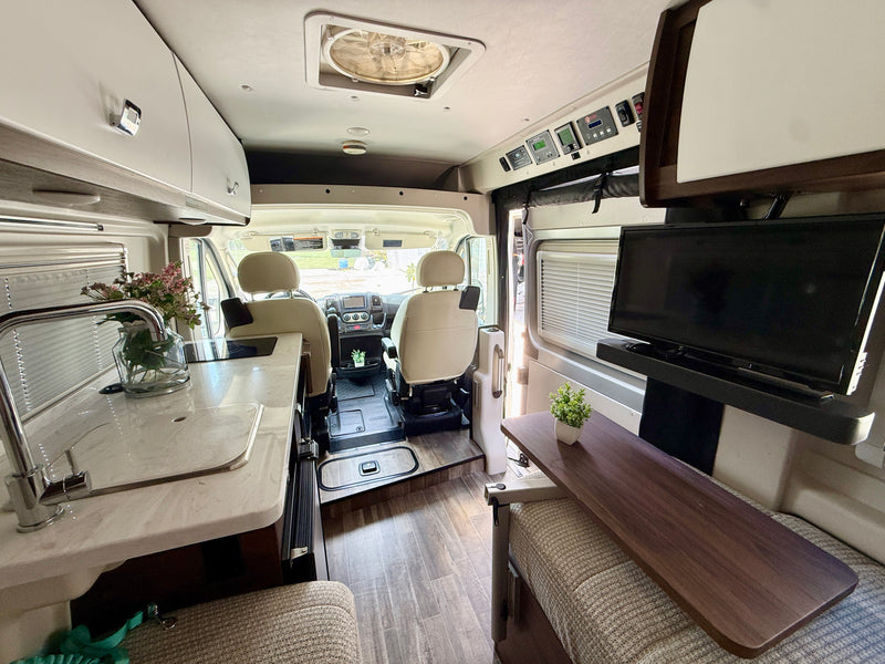 View inside a modern rv rental showing a kitchen counter with sink, swivel front seats, overhead storage, and a lounge with TV. — photo 15