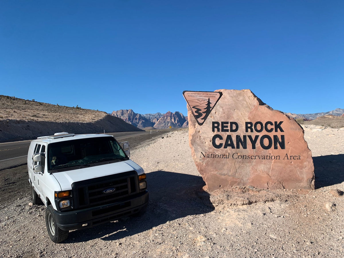 A white Ford van RV rental parked by the Red Rock Canyon National Conservation Area sign
