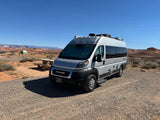 A silver rv rental camper van is parked at a sunny desert campsite with a picnic table and red rock mesas under a clear blue sky.