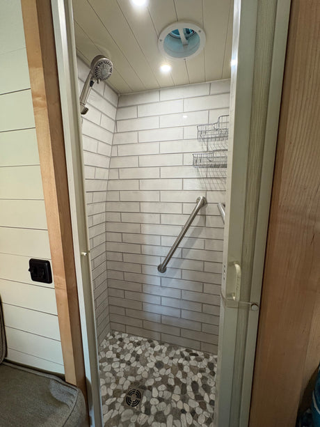 RV rental shower with sleek subway tile walls, pebble floor, grab bar, storage, and ceiling vent.
