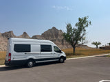 White RV rental parked on asphalt, facing the unique geological formations of the Badlands, with dry grass & a tree.
