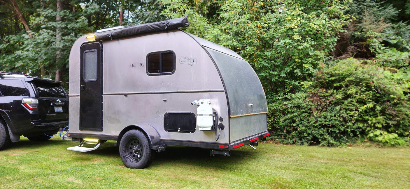 A silver teardrop rv rental trailer is hitched to a black SUV on grass, perfect for camping trips.