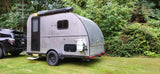 A silver teardrop rv rental trailer is hitched to a black SUV on grass, perfect for camping trips.