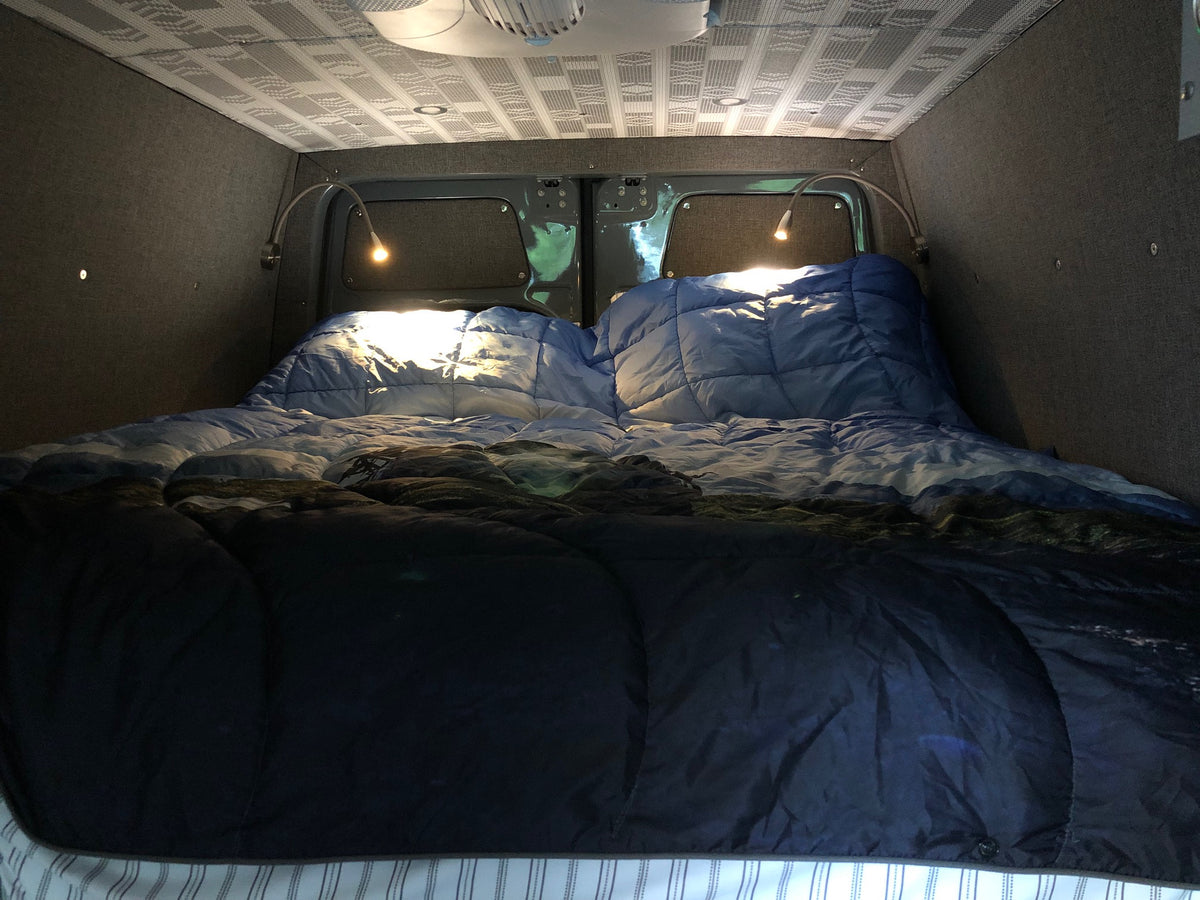 Cozy RV rental interior: A comfortable bed with blue comforters and two bright reading lights. Custom walls and ceiling. — photo 12