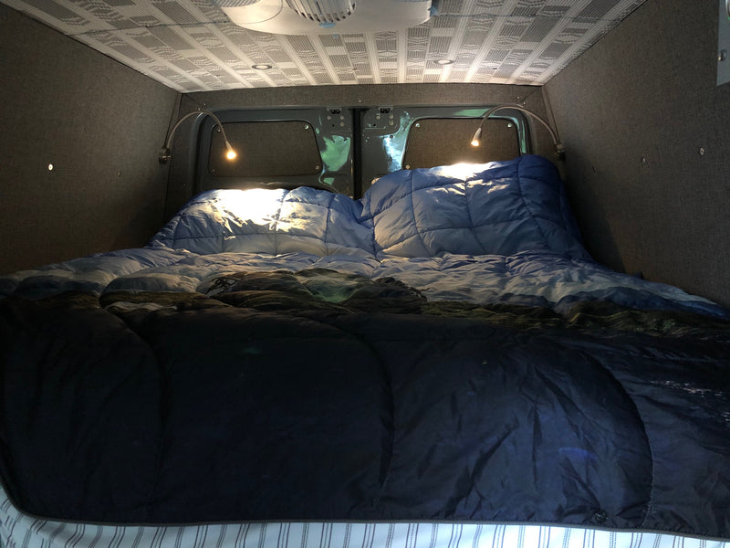 Cozy RV rental interior: A comfortable bed with blue comforters and two bright reading lights. Custom walls and ceiling. — photo 12