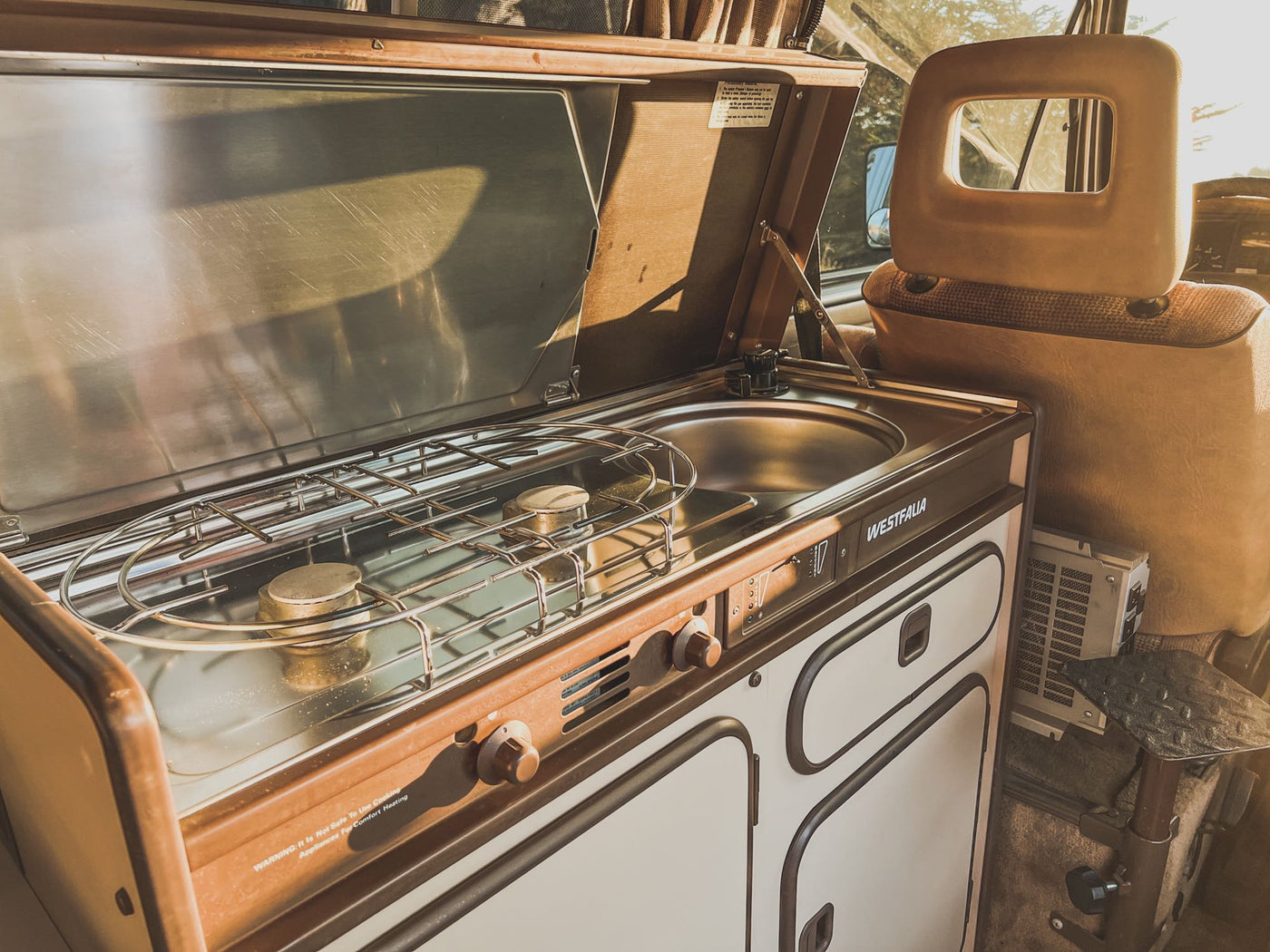  watching a golden ocean sunset from inside a pop-up camper.|Cozy Westfalia rv rental interior showing a vintage kitchenette with stove and sink bathed in warm sunlight — photo 10