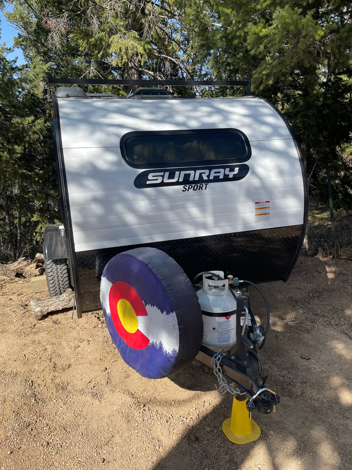 Compact Sunray Sport teardrop camper rv rental, featuring a Colorado flag tire cover & propane tank, nestled in the woods.