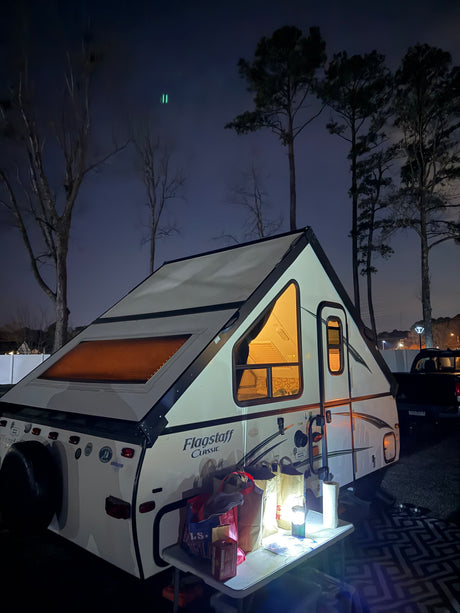 Nighttime rv rental scene featuring an illuminated A-frame camper, outdoor table, and trees under a dark sky.