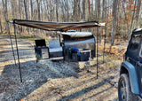 Compact teardrop RV rental with an extended awning and outdoor kitchen setup at a wooded campsite.