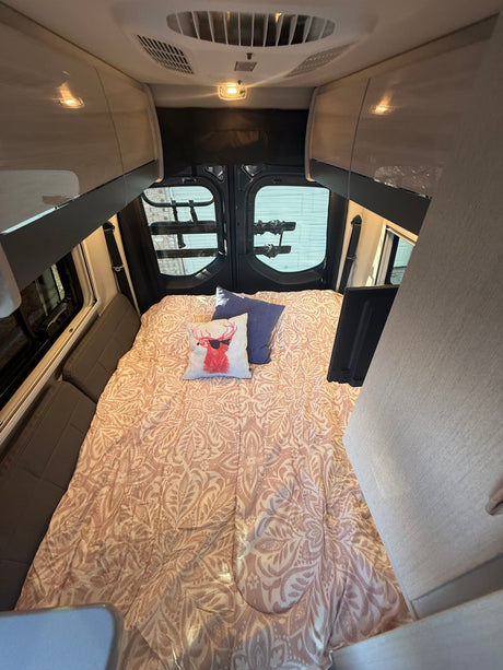 Cozy RV rental bedroom with patterned comforter, two pillows, overhead cabinets, and rear windows.
