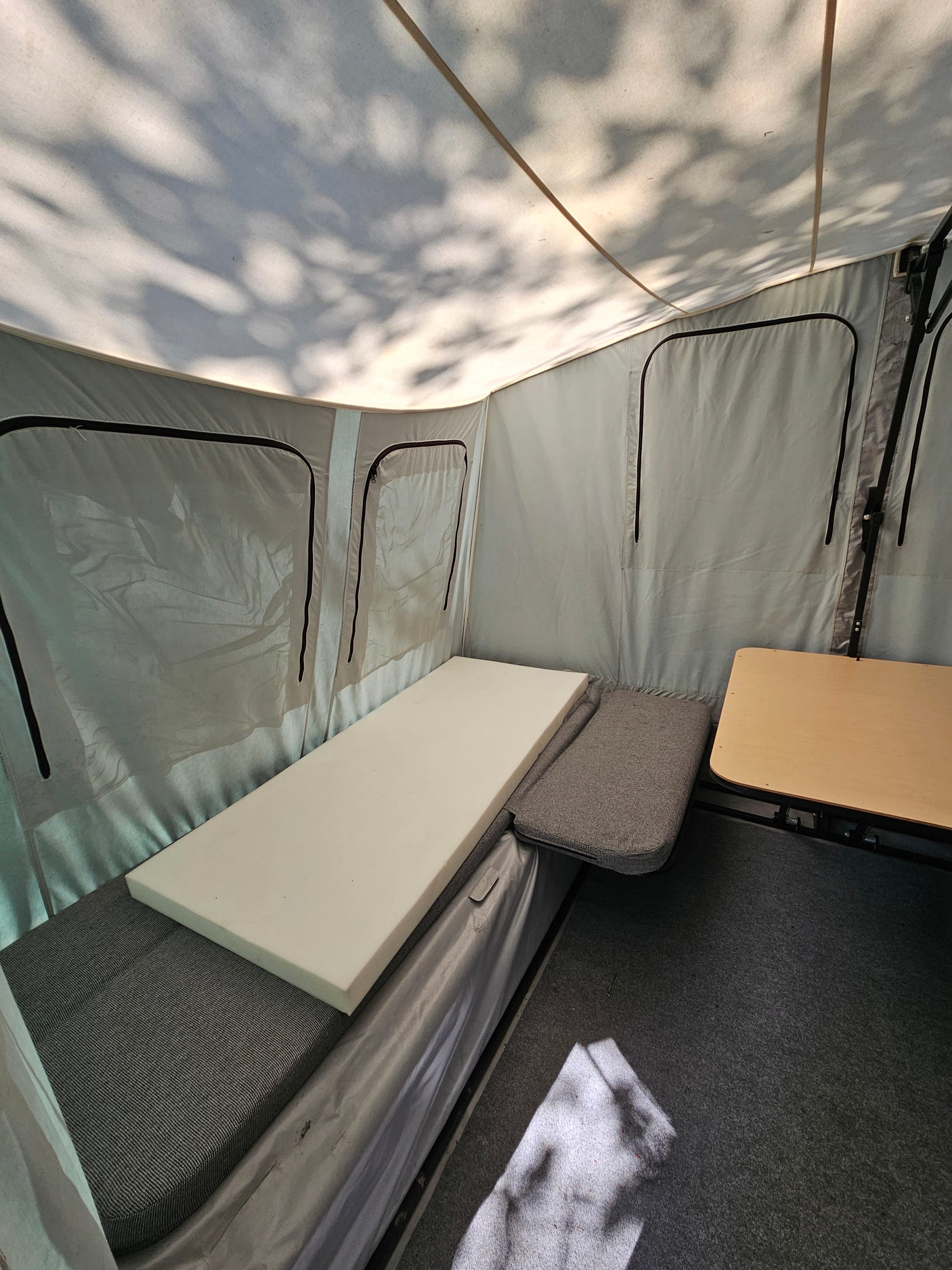 Inside an RV rental pop-up camper: fabric bed with white foam, small table, and dappled shadows on the canvas ceiling.