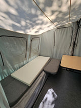 Inside an RV rental pop-up camper: fabric bed with white foam, small table, and dappled shadows on the canvas ceiling.