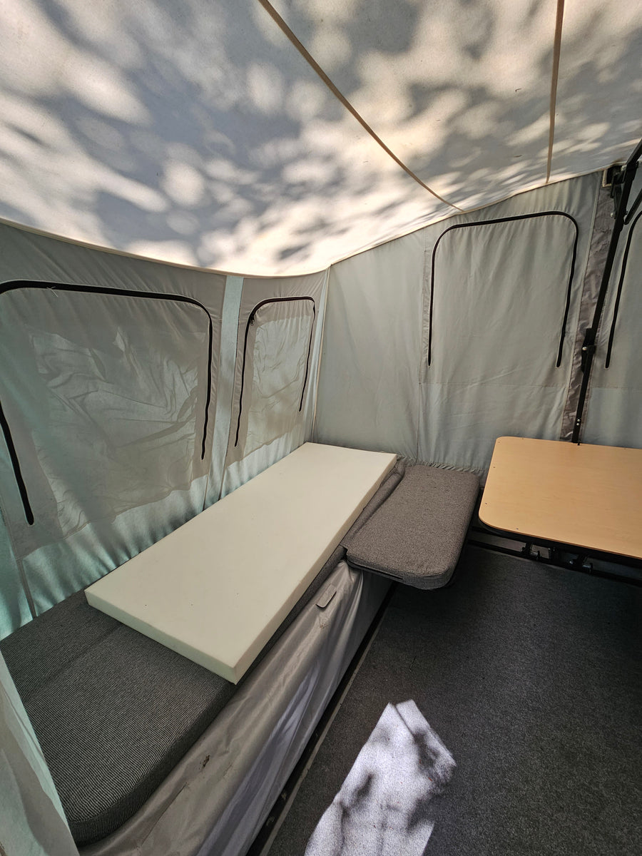 Inside an RV rental pop-up camper: fabric bed with white foam, small table, and dappled shadows on the canvas ceiling.