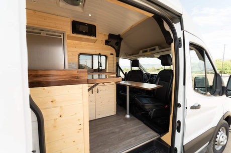 Explore this modern RV rental's cozy, wood-paneled camper van interior with a kitchen, dining table, and front seats, ready for travel.