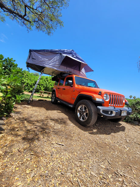 An orange Jeep with a rooftop tent, ideal for an RV rental, parked in a scenic outdoor camping spot under a blue sky.