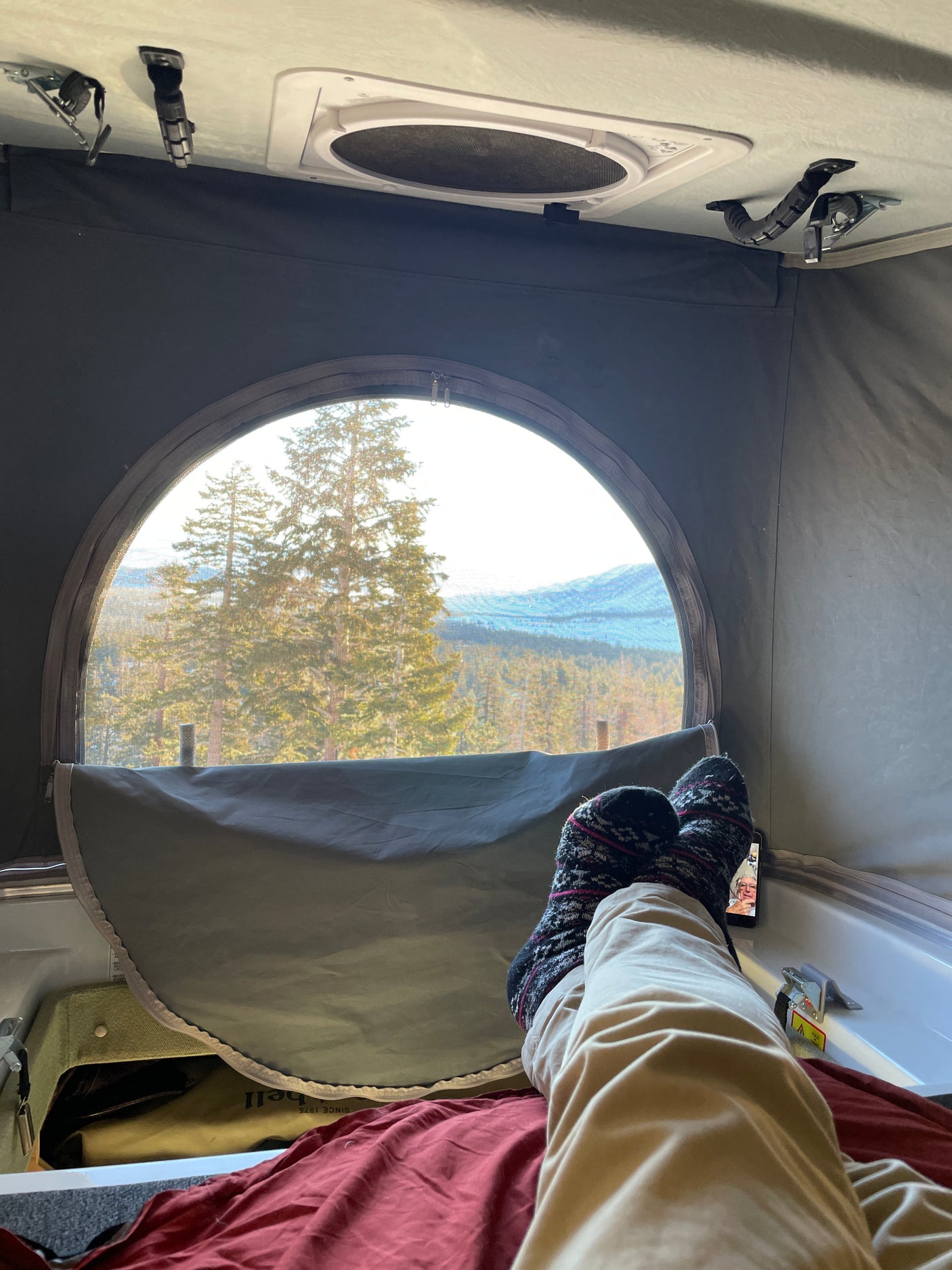 A cozy RV rental experience: Legs in socks, phone nearby, with a beautiful mountain forest view from the round window.