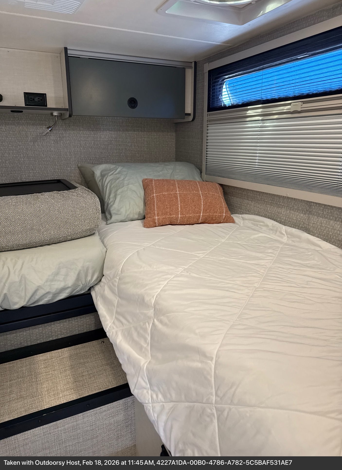 Cozy RV rental bedroom featuring a comfortable bed with soft bedding, pillows, and a window with blinds showing bright blue sky.