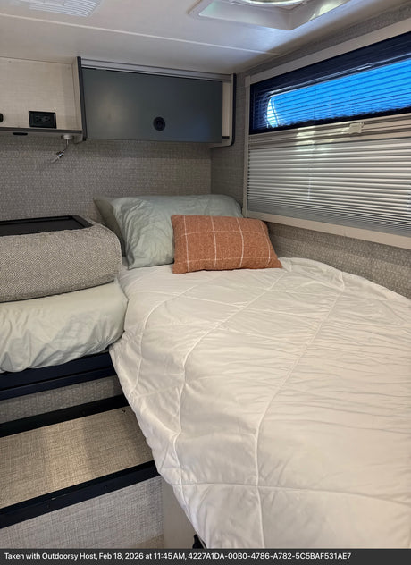 Cozy RV rental bedroom featuring a comfortable bed with soft bedding, pillows, and a window with blinds showing bright blue sky.