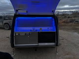 Teardrop RV rental: Illuminated outdoor kitchen with sink, stove, and storage. Night camp scene with snowy mountains.