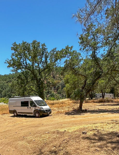 White camper van (rv rental) parked on a dirt camping spot in a sunny, tree-filled outdoor area with a clear blue sky.