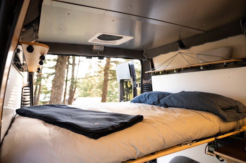 An inviting RV rental sleeping area: bed with light sheets, dark pillows/blanket, and forest views outside. — photo 7