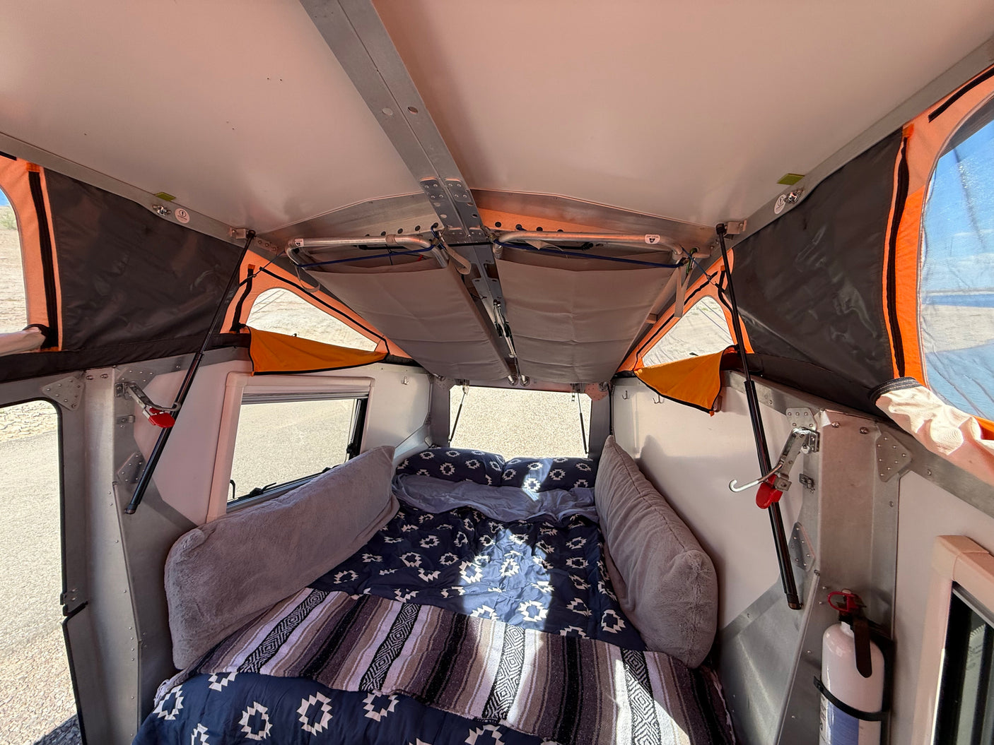 Inside an RV rental pop-up camper, showcasing a cozy bed with blankets, overhead storage, and a fire extinguisher.