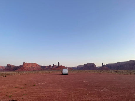 An RV rental parked in a vast, red desert with towering sandstone formations under a clear blue sky, perfect for adventure.