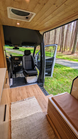 RV rental van interior showing wood ceiling, floor, and swivel seats, with the side door open to a scenic forest path.