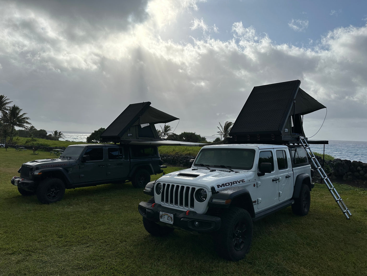 Two Jeep Gladiator RV rentals with rooftop tents by the ocean. Perfect for an off-road camping adventure on grassy terrain.