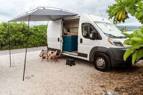 White camper van RV rental with awning, chairs, and custom interior, set up on sand for beachside camping.
