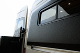 Side view of an RV rental: dark textured panel with an open window revealing the interior. Rear door and mounting bracket visible.