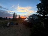 RV rental at a stunning sunset campsite. Camper van, chairs, and trees silhouetted under a vibrant, colorful sky.