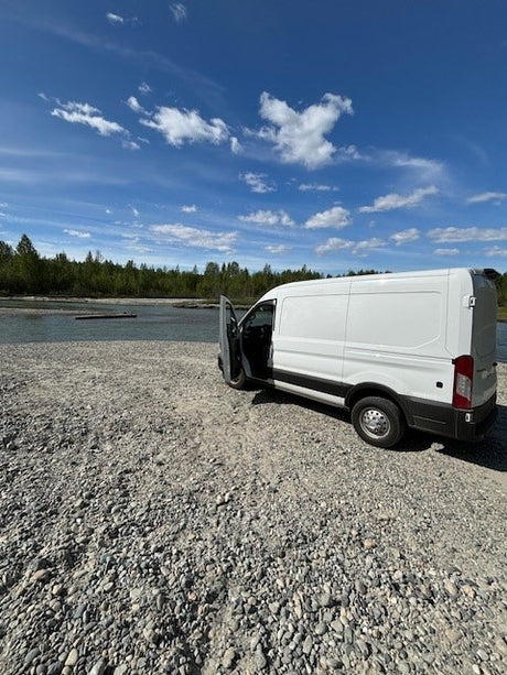 Scenic RV rental van stop: white van on a gravel riverbank with trees, water, and a bright blue, cloudy sky.