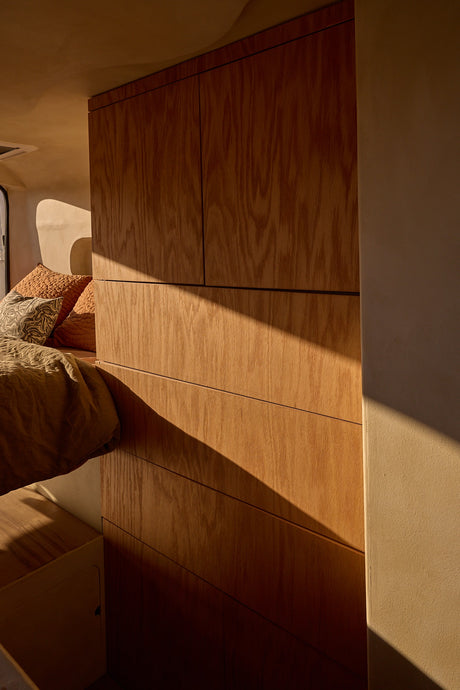 Sunlit RV rental interior with minimalist wood cabinets & cozy bed. Warm light casts diagonal shadows.