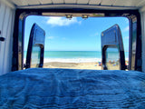Waking up to this ocean view from an RV rental bed. Open van doors frame a serene beach scene.