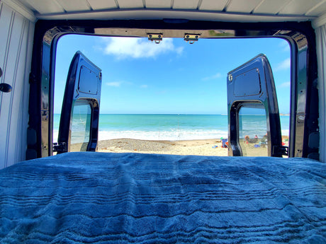 Waking up to this ocean view from an RV rental bed. Open van doors frame a serene beach scene.