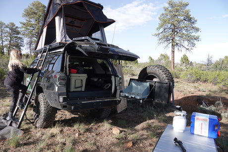 Enjoy an RV rental adventure! Woman accessing a rooftop tent on an SUV, equipped for camping with a stove and gear.