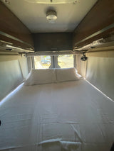 RV rental sleeping area: a neatly made bed with white sheets & pillows, overhead cabinets, and back windows.