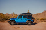 A bright blue Jeep Wrangler with a rooftop tent, resembling an RV rental, on a desert trail among saguaro cacti and mountains.