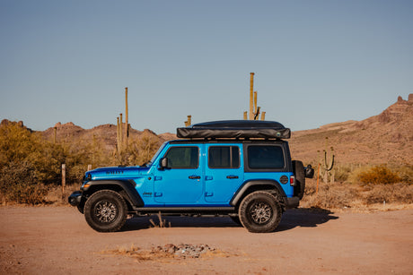 A bright blue Jeep Wrangler with a rooftop tent, resembling an RV rental, on a desert trail among saguaro cacti and mountains.