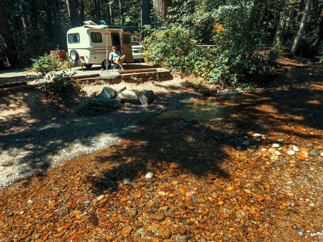 Man relaxing by a clear forest creek with his RV rental at a serene campsite.