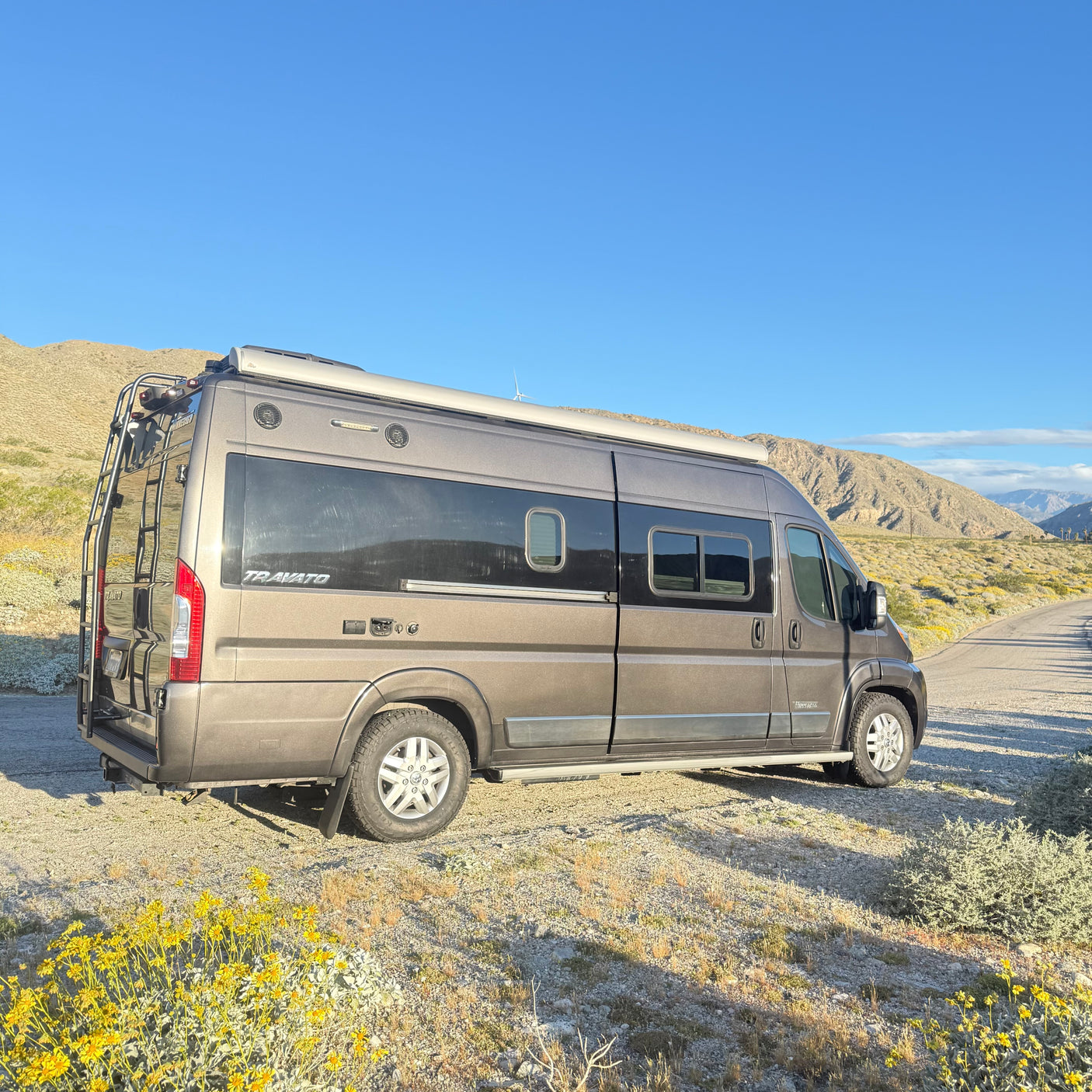 Gray Winnebago Travato **RV rental** parked on a dirt road with yellow flowers, mountains, and a clear blue sky.