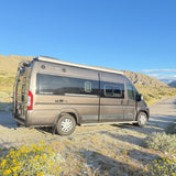 Gray Winnebago Travato **RV rental** parked on a dirt road with yellow flowers, mountains, and a clear blue sky.