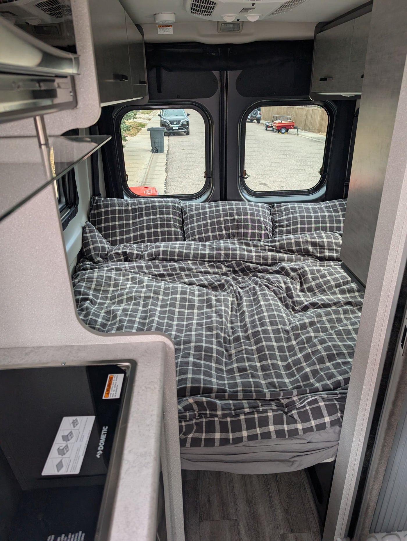Inside a compact RV rental: a comfy bed with plaid bedding, overhead cabinets, and a street view through the rear windows. — photo 4
