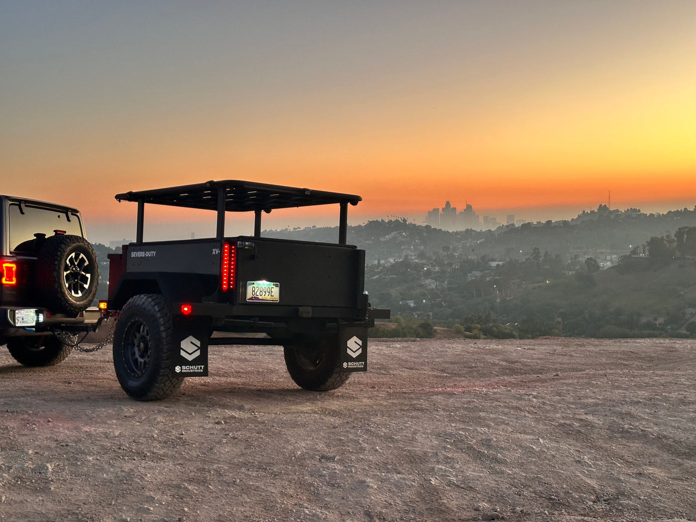 A dark Jeep tows a rugged Schutt Industries overland trailer at sunset, overlooking a hazy city. Ideal for your next RV rental adventure. — photo 10